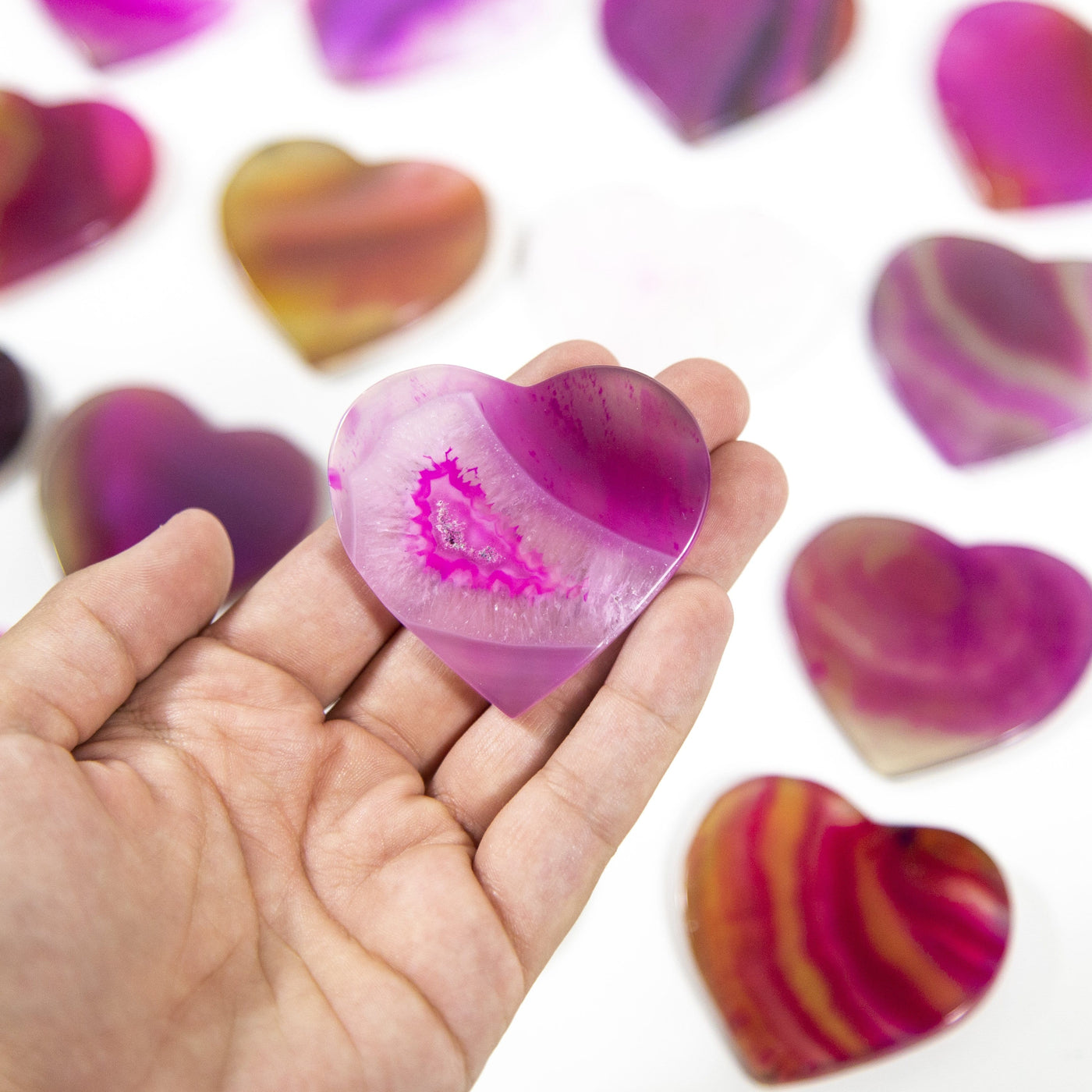 A pink Agate Heart Shaped Cabochon in a hand, multiple agate hearts in the background on a white surface.
