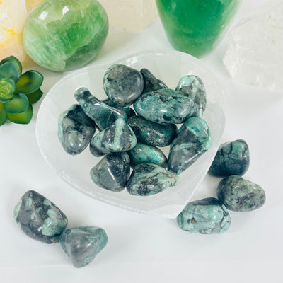 Emerald Crystal Large Tumbled Stone - By the Piece several in bowl with several outside of bowl with props and plants in the background