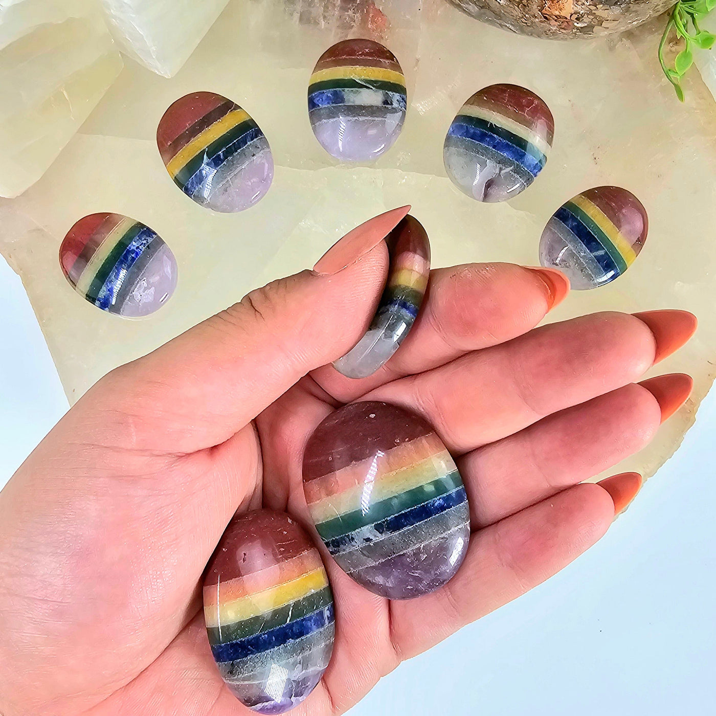 Seven Chakra Worry Stone three rainbow colored stones in hand, one between index finger and thumb, with 5 other rainbow colored 7 chakra stones in background on a quartz platter