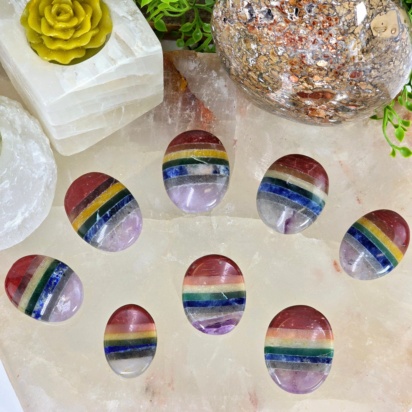 Seven Chakra Worry Stone 5 rainbow colored stones arranged face up and 3 rainbow colored stones below with indentation side up with props and plants in the background