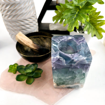 Fluorite Crystal Candle Holder - AA Grade one placed next to bowl with palo santo on rose quartz platter with plants and props in the background