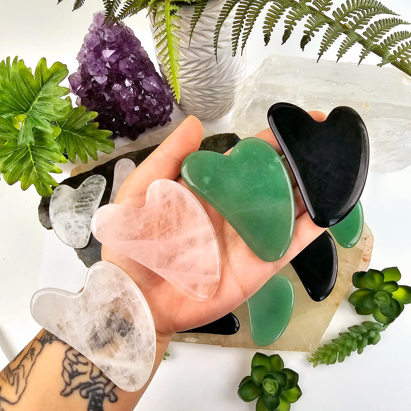 Stone Gua Sha Face Massage Tool all variants in hand with more variants in the background with props and plants