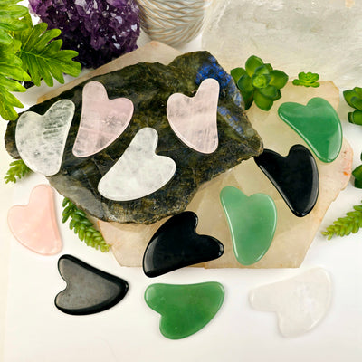 Stone Gua Sha Face Massage Tool - all variants arranged on stone platters with props and plants in the background