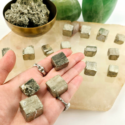 Pyrite Cube - You Choose - 4 variants in hand with other variants in background on quartz platter with plants and props