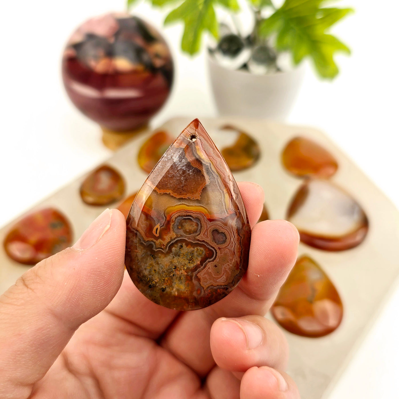 Carnelian Teardrop Crystal Pendant - You Choose - variant 10 in hand for size reference with other variant in the background on stone platter with props and plants in the background