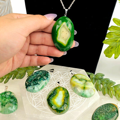 Agate Druzy Crystal Pendant - Dyed Green - one on chain on black necklace stand with hand for size reference with others on selenite coaster and log with plants in the background