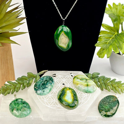 Agate Druzy Crystal Pendant - Dyed Green - six different pendants, one on chain on black necklace stand, five on selenite coaster and log with plants in the background