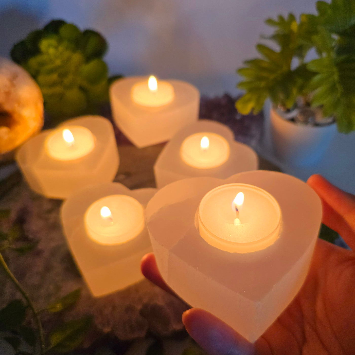 Smaller Selenite Heart Shaped Candle Holder one candle holder in hand with lit candle for size reference with other candle holders with lit candles in the background with props and plants in dim lighting