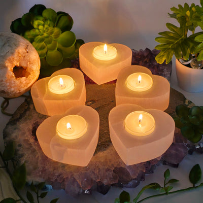 Smaller Selenite Heart Shaped Candle Holder five heart candle holders with lit candles on amethyst stone platter with props and plants in the background in dim lighting