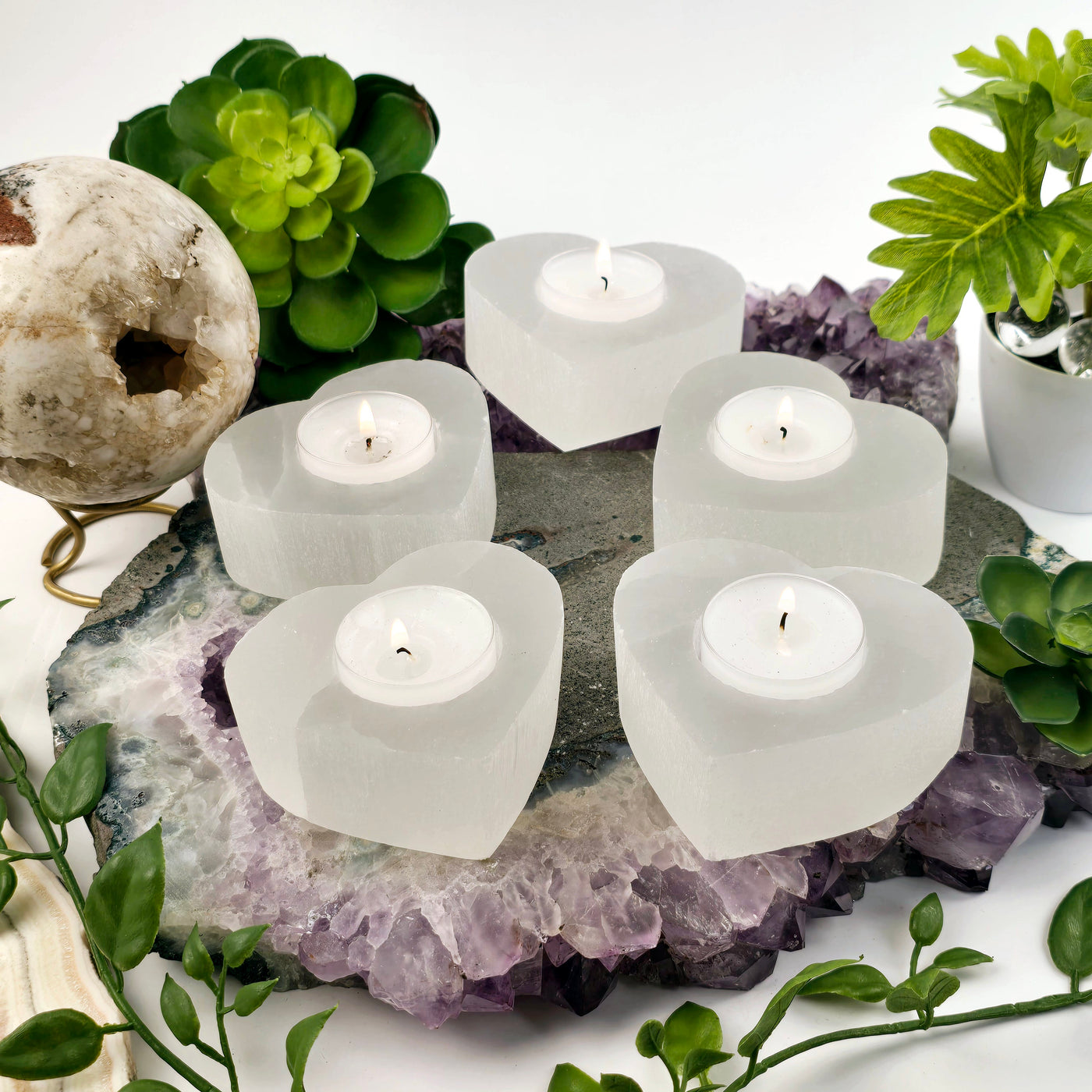 Smaller Selenite Heart Shaped Candle Holder - five candle holders with lit candles in bright lighting on stone platter with plants and props in the background