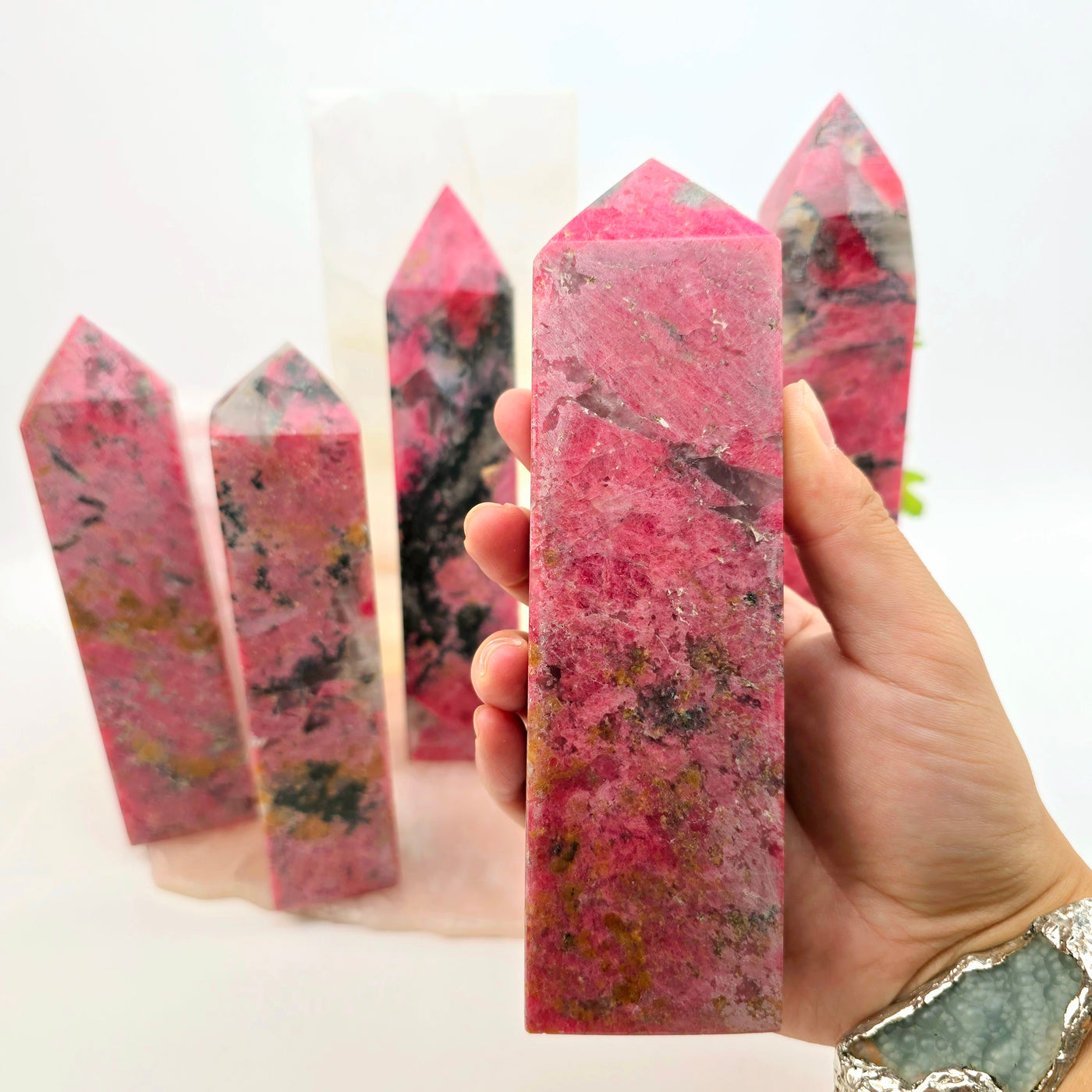 Rhodonite Polished Crystal Tower - You Choose - variant 2 in hand for size reference with other variants in background with props and plants