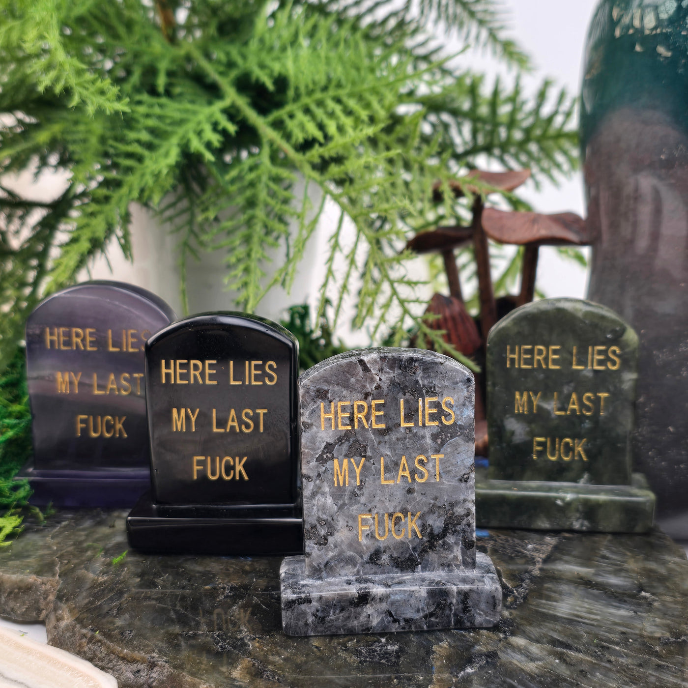 Crystal Gravestone with Engraved "Here Lies My Last F***" - You Choose Stone - Purple Fluorite, Black Obsidian, Larvikite, and Nephrite Jade variants in row on stone platter with props and plants in the background