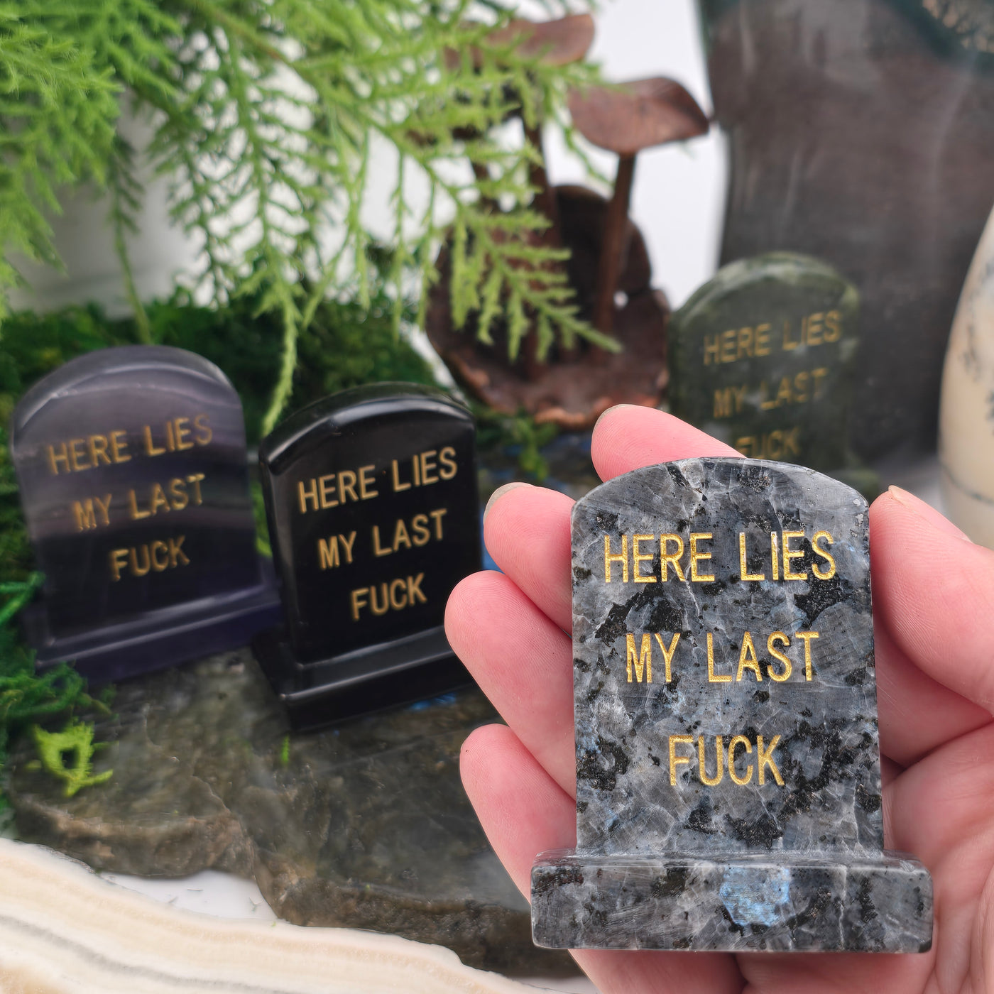 Crystal Gravestone with Engraved "Here Lies My Last F***" - You Choose Stone - Larvikite variant in hand for size reference with other variants in background on stone platter with props and plants