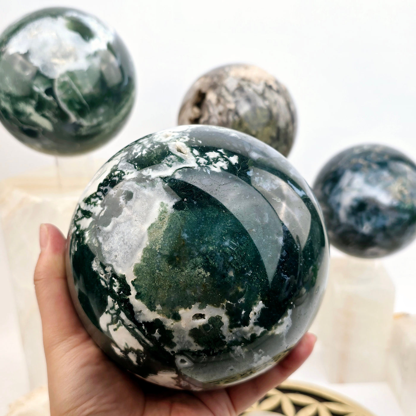 Moss Agate Large Crystal Spheres - You Choose - variant 4 in hand with other variants in background on crystal pedestals and acrylic sphere stands