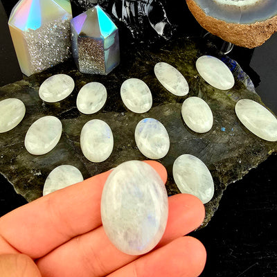 Moonstone Oval Crystal Cabochon - You Choose - one in hand for size reference with other variants on labradorite platter on black background with props