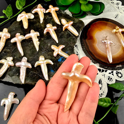 Cross Shaped Freshwater Pearl - Undrilled - You Choose - variant 20 in hand for size reference with other variants on labradorite platter, black acrylic, and natural agate slice over a white doily with plants in the background