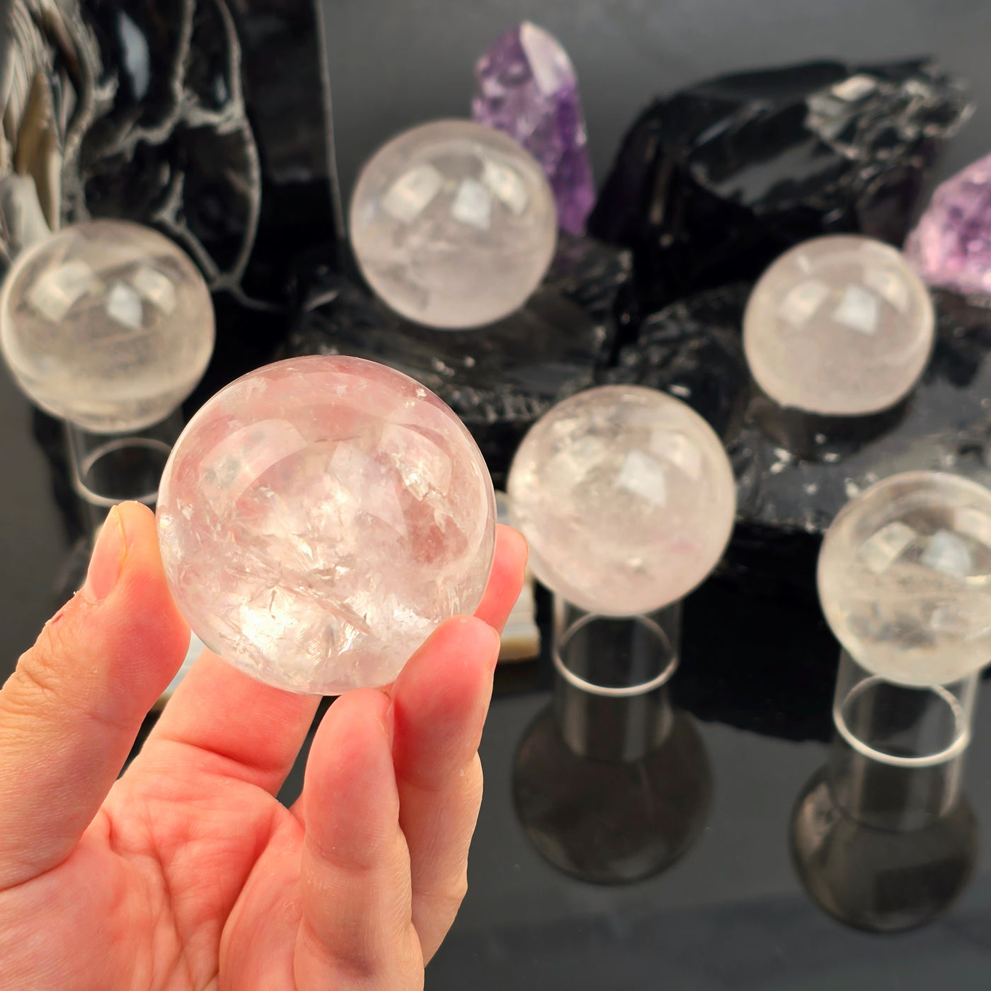 Rose de France Amethyst Crystal Sphere - You Choose - one variant in hand for size reference with other variants in background on black acrylic and black obsidian sphere holders with amethyst points