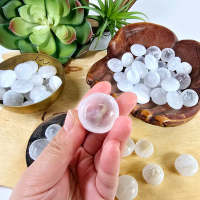 Optical Selenite Crystal Tumbled Stone - By the Piece - one in hand to show size reference and clarity with others in bowls and dishes and on wooden platter with succulents and props in the background