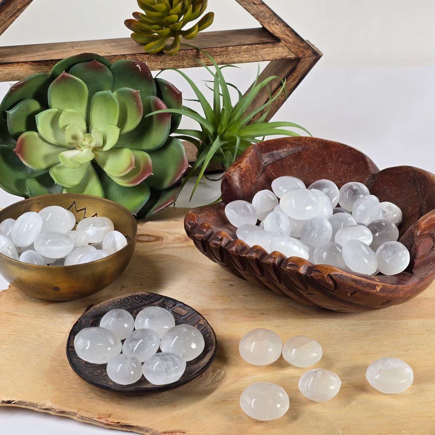 Optical Selenite Crystal Tumbled Stone - By the Piece - stones in various bowls and dishes and on wooden platter with shelf and succulents in the background