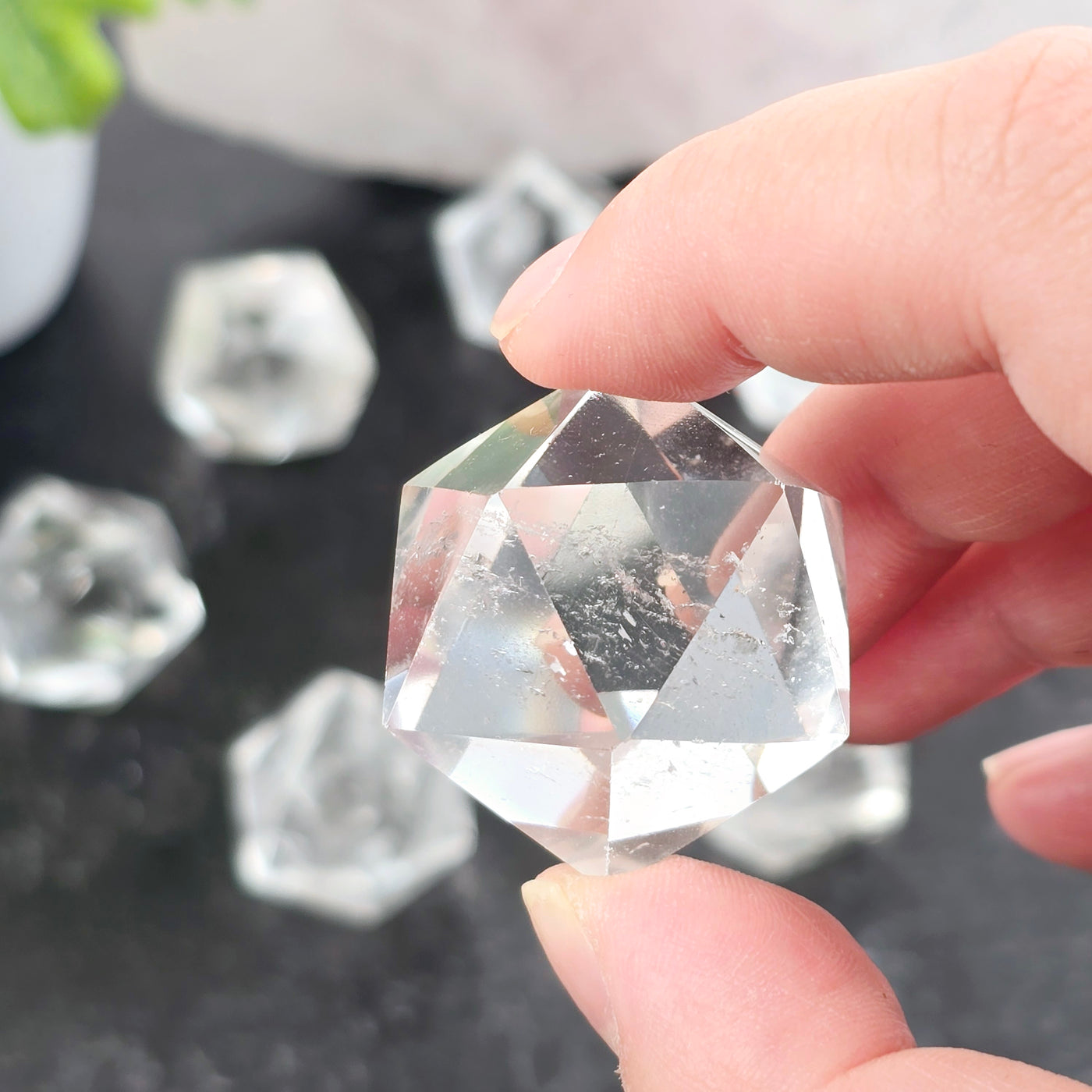 Crystal Quartz Small Icosahedron - one in hand for size reference with others in the background on dark table with props and plants