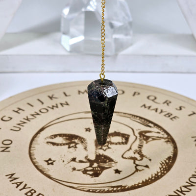 Garnet Crystal Pendulum with Gold Plated Bail and Chain - one hanging over a pendulum board with a crystal in the background on a white triangular shelf