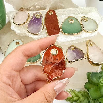 Crystal Freeform Slice Pendant with 24k Gold Electroplated Edges Top Front Drilled - You Choose Stone - red jasper variant in hand with other variants in the  background on stone platters with props and plants