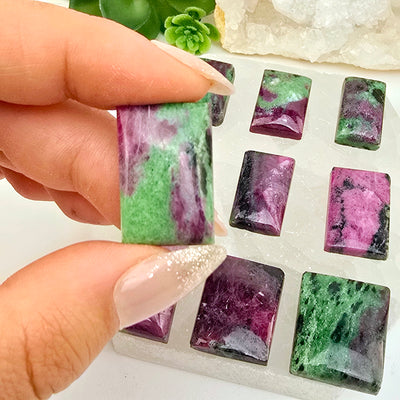 Ruby Zoisite - Anyolite - Crystal Rectangle Cabochon - You Choose variant 4 in hand for size reference with other variants in the background on selenite platter with plants and props