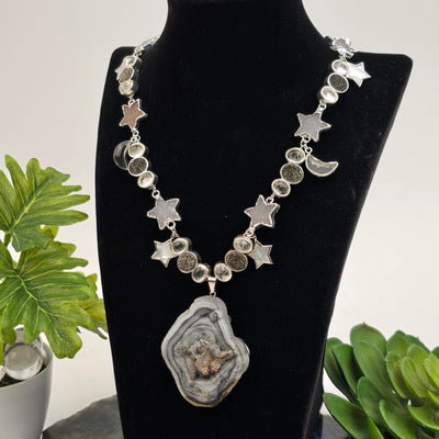 Chalcedony Druzy Galaxy Pendant Sterling Silver Necklace with Druzy and Mother-of-Pearl Stars and Crystal Quartz Moon Charms - Handmade - One-of-a-Kind on necklace stand with plants in the background