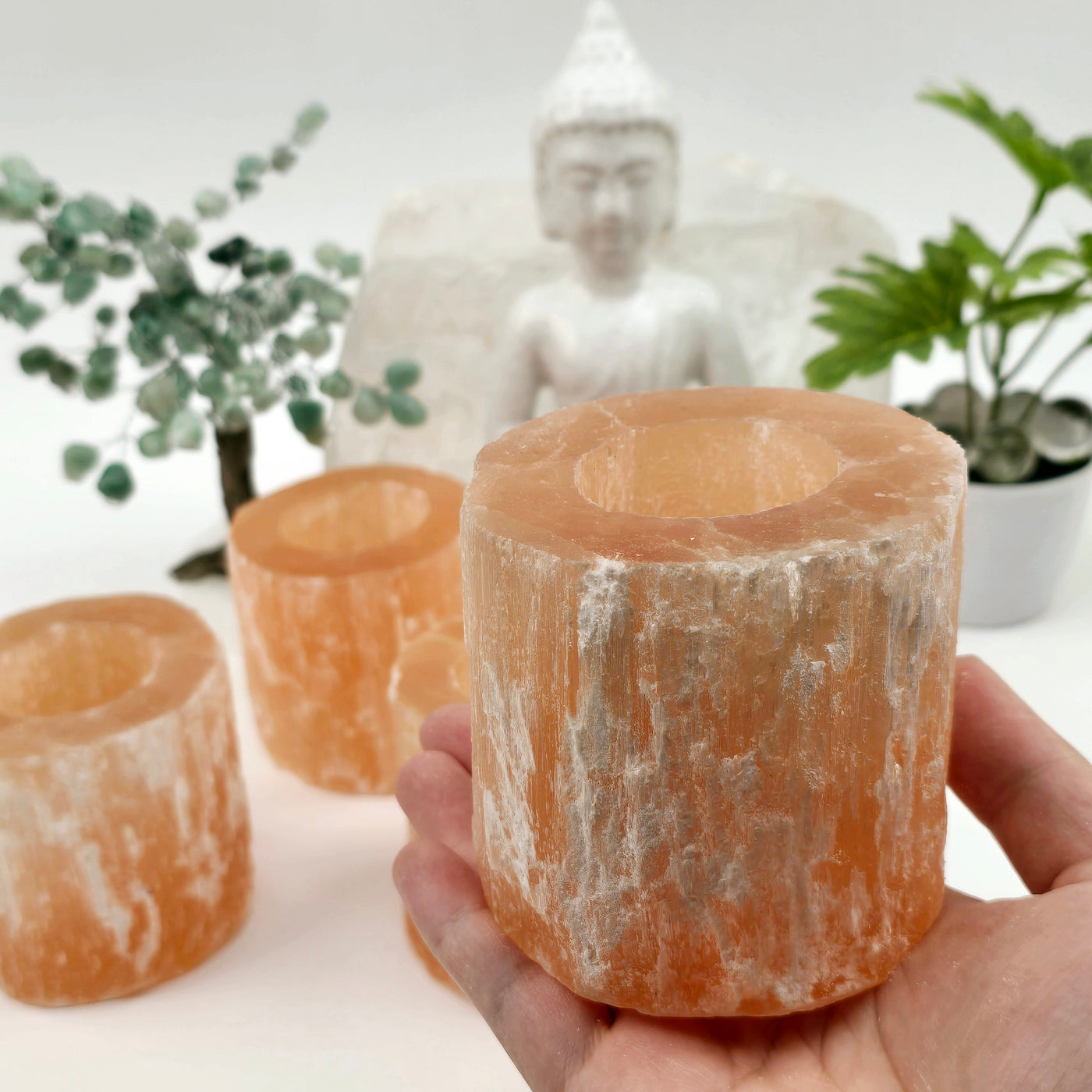 Orange Selenite Crystal Candle Holder one in hand in bright lighting for size reference with others in the background with props and plants