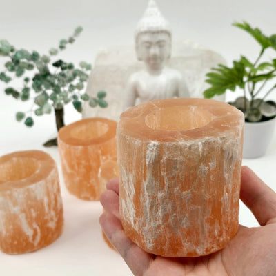 Orange Selenite Crystal Candle Holder one in hand in bright lighting for size reference with others in the background with props and plants