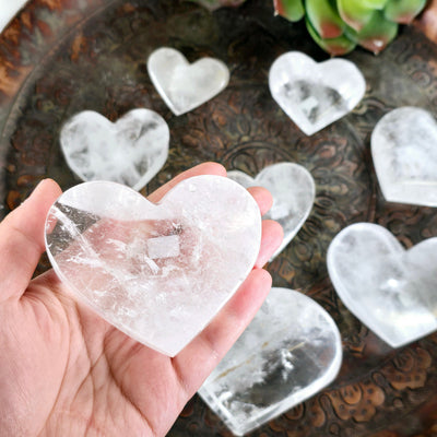 Crystal Quartz Polished Heart - You Choose #2 one variant in hand for size reference with other variants in the background with props and plants