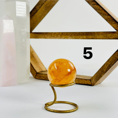 Golden Healer Crystal Sphere labeled variant 5 on a metal stand on a white table with a wood shelf and two crystal points in the background