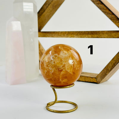 Golden Healer Crystal Sphere labeled variant 1 on a metal stand on a white table with a wood shelf and two crystal points in the background