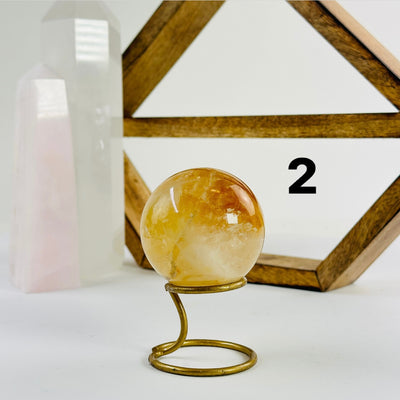 Golden Healer Crystal Sphere labeled variant 2 on a metal stand on a white table with a wood shelf and two crystal points in the background 