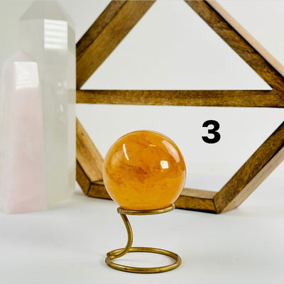 Golden Healer Crystal Sphere labeled variant 3 on a metal stand on a white table with a wood shelf and two crystal points in the background