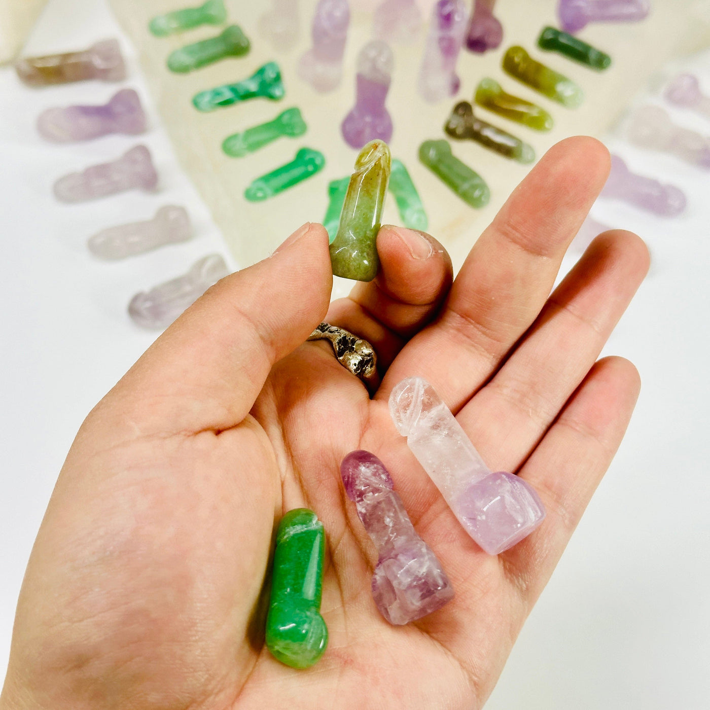 Mini Gemstone Penis - Carved Penis - green aventurine mini crystal penis between fingers with one green aventurine and two amethyst mini crystal penises in hand with other crystal penises arranged in the background