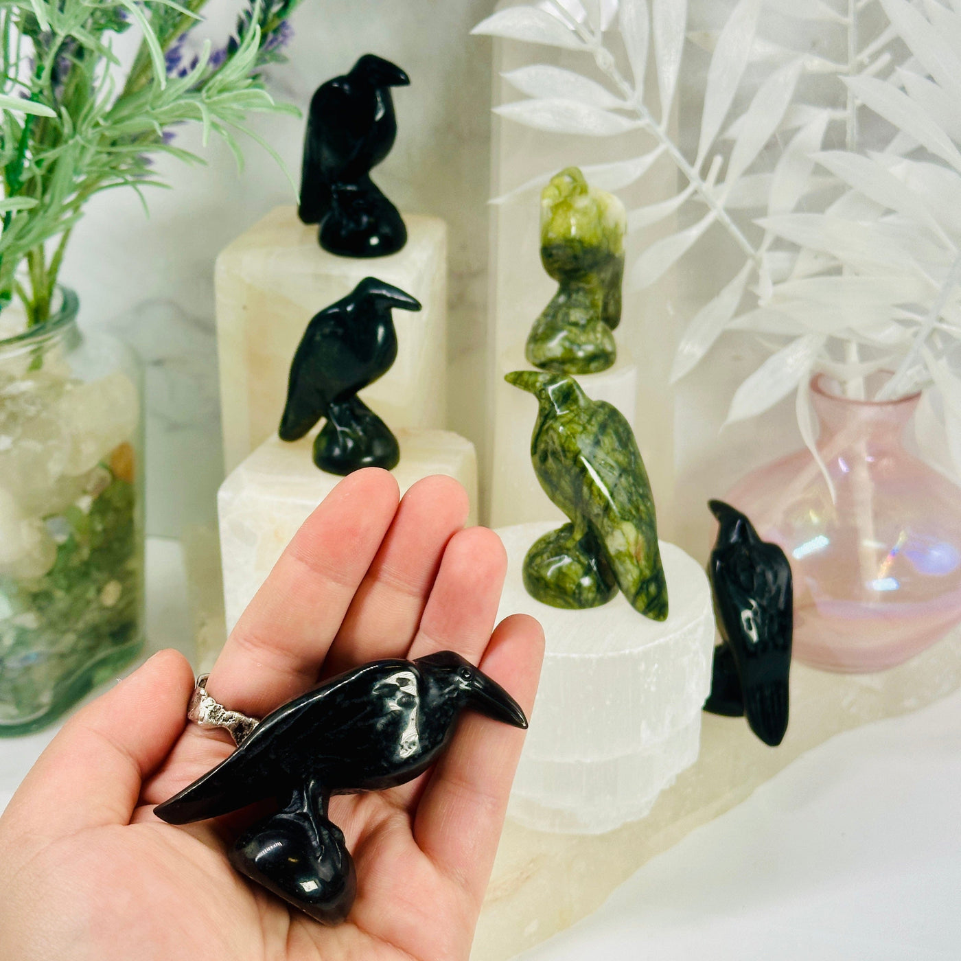 Gemstone Raven - Carved Raven - black obsidian carved raven in hand for size reference with other crystal carved ravens in the background on white pedestals of different heights