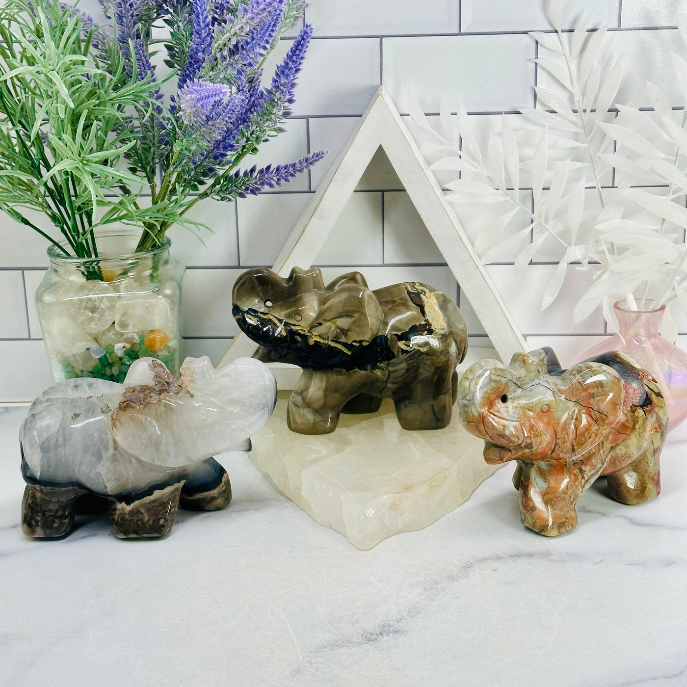 Lava Agate Crystal Carved Elephants - YOU CHOOSE all elephant variants at different angles arranged on marble textured ground and white brick pattern wall with props in backround