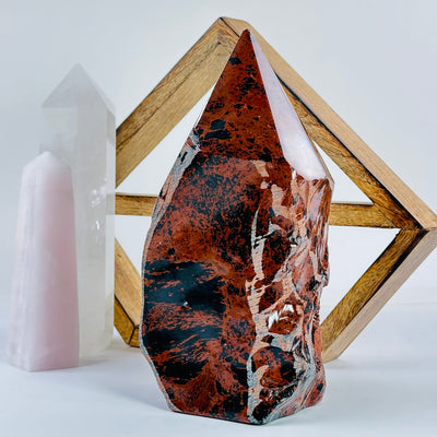 Mahogany Obsidian Crystal Semi Polished Point front view on a white table with a wood shelf and 2 crystal points in the background.