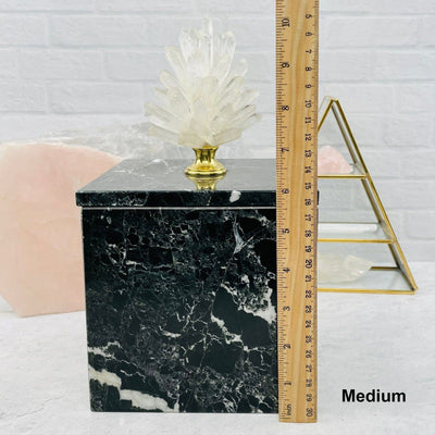 medium crystal point pinecone on marble box  next to a ruler for size reference with decorations in the background