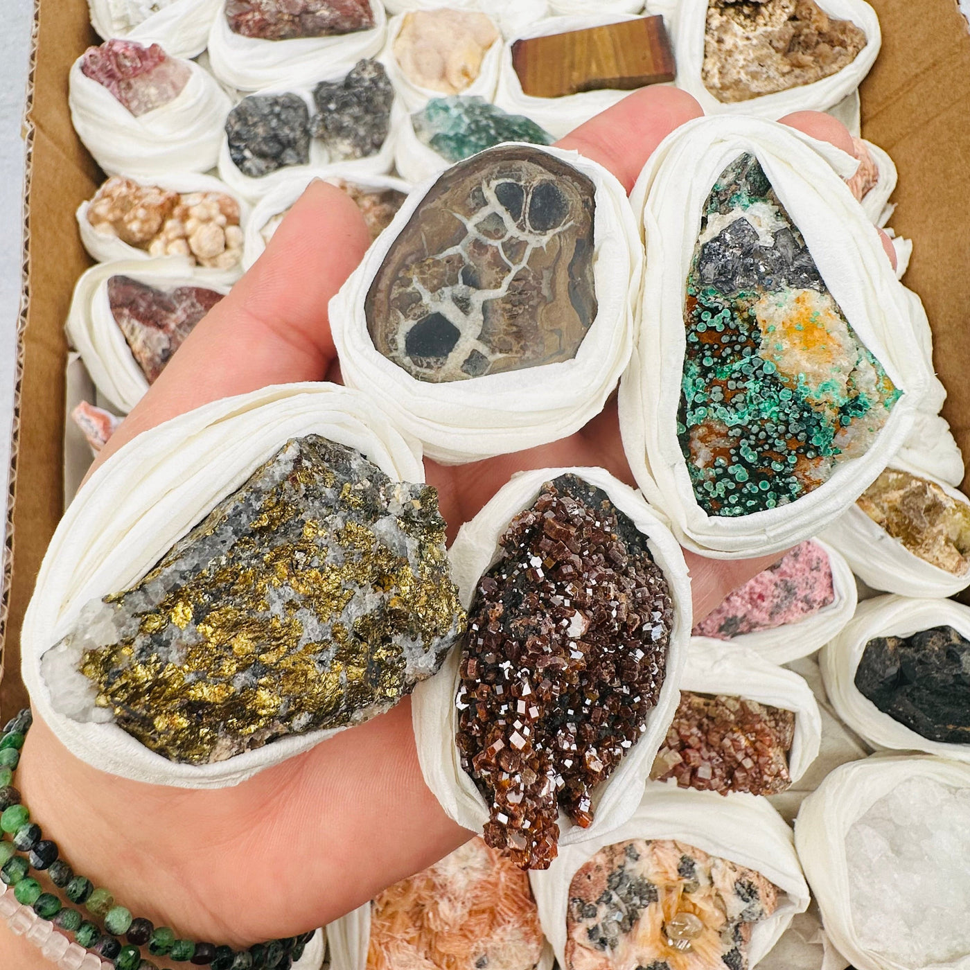 Assorted minerals in a woman's hand with more in a box underneath.