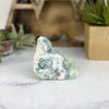 Raw Fluorite Crystal Stone on marble base with green plants in background.