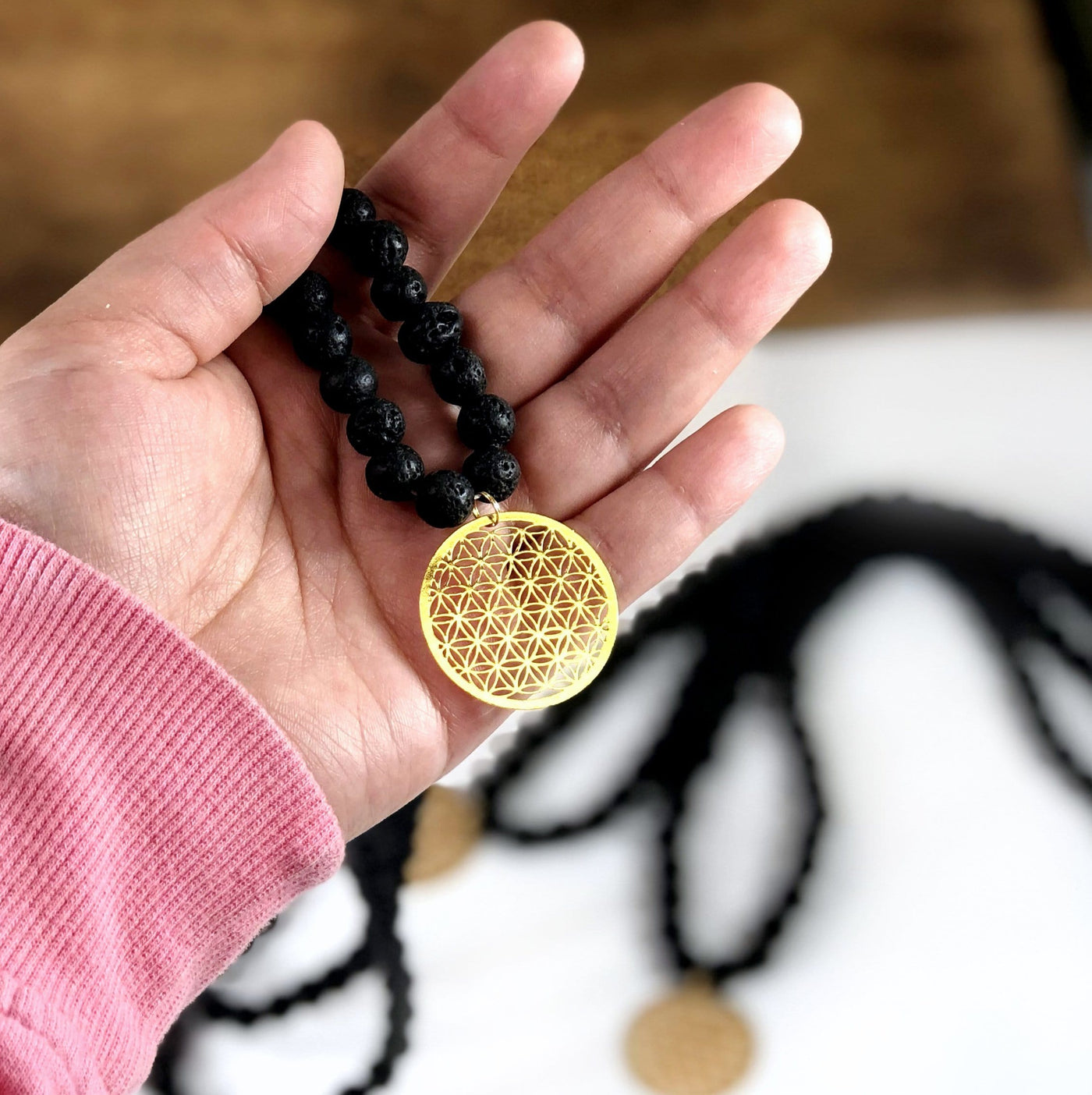 Diffuser Necklace Lava Bead with Flower of Life Charm in Gold in Hand on White Background.