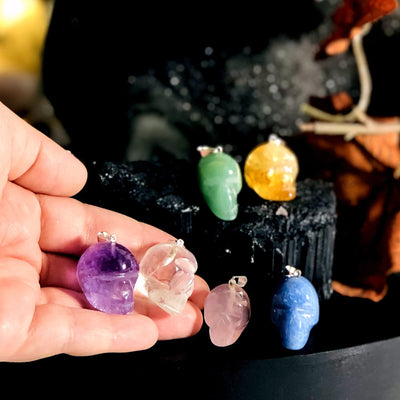 two skull shaped gemstone pendants in hand for size reference with four others in background display