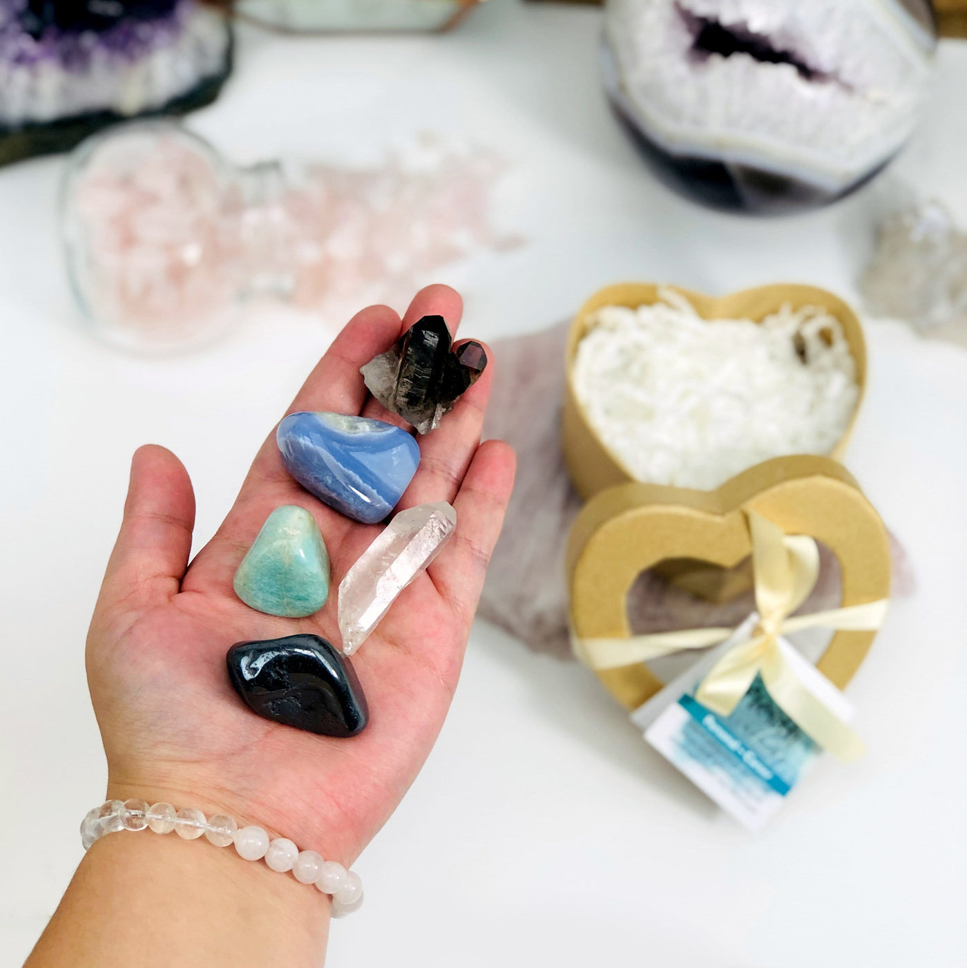 hand holding up smoky quartz, blue lace agate, amazonite, crystal quartz, and hematite with growth heart box blurred in the background with decorations