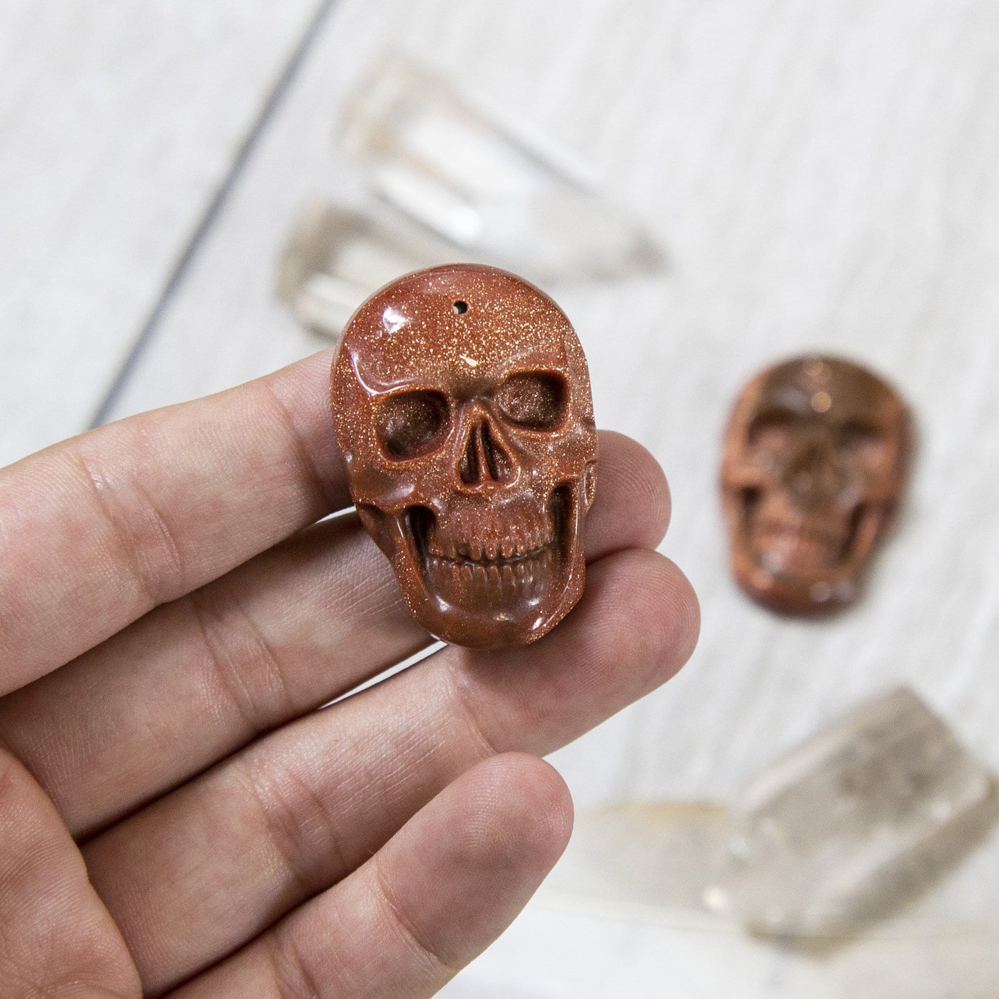 one skull shaped goldstone piece in hand for size reference with one other in background display