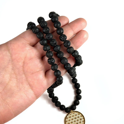 Close Up of Diffuser Necklace Lava Bead with Flower of Life Charm in Gold in Hand on White Background.