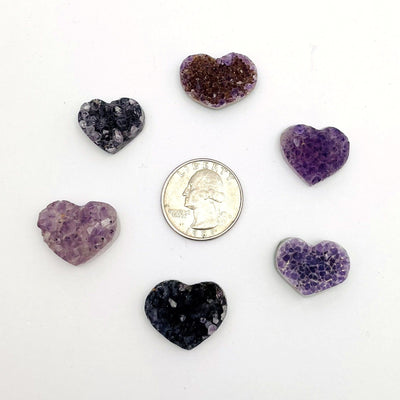 Assorted shades of amethyst hearts in light purple, dark purple, black, purple with brown and other variety on a white background with a quarter for sizing reference.