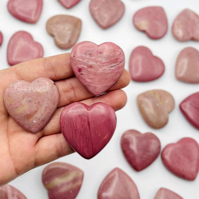 Hand holding up 3 Rhodonite Heart Shaped Stones with more scattered in the background