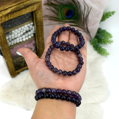 hand holding 2 Amethyst Round Bead Bracelets while wearing 2 others with decorations in the background